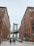 A city street view featuring the Manhattan Bridge framed by two large brick buildings. People walk along the cobblestone street, and cars are parked along the sides. The scene captures an urban atmosphere with architectural details and the iconic bridge in the background.