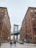 A city street view featuring the Manhattan Bridge framed by two large brick buildings. People walk along the cobblestone street, and cars are parked along the sides. The scene captures an urban atmosphere with architectural details and the iconic bridge in the background.