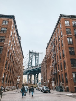 A city street view featuring the Manhattan Bridge framed by two large brick buildings. People walk along the cobblestone street, and cars are parked along the sides. The scene captures an urban atmosphere with architectural details and the iconic bridge in the background.