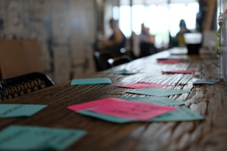 A long wooden table with several colorful post-it notes scattered across its surface. The focus is on the notes in the foreground, while the background blurs into people sitting and working in what appears to be a cafe or co-working space.