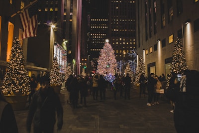 A bustling city street at night adorned with festive Christmas decorations. Large, illuminated Christmas trees line the pathway, surrounded by crowds of people enjoying the holiday atmosphere. Many are engaged in activities such as taking photos, walking, and admiring the lights. The urban backdrop is composed of tall buildings with additional lights and decorations.