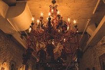 A large, decorative chandelier draped in red beads and illuminated by warm, glowing lights. The setting is indoors with exposed brick walls and a wooden ceiling. Several Santa Claus figures are hanging or attached to the chandelier, creating a festive, holiday atmosphere.
