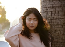 A young woman with long dark hair stands beside a textured tree trunk, wearing a light-colored sweater and looking into the camera. The lighting creates a warm, golden glow, suggesting it is during sunset. The background is softly blurred, with hints of greenery and water.