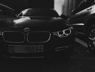 A close-up photograph of a luxury car front, featuring prominently its distinctive grille and headlights. The setting appears to be an urban outdoor environment with other vehicles parked nearby. The image is in black and white, creating a dramatic and moody effect.