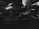 A close-up photograph of a luxury car front, featuring prominently its distinctive grille and headlights. The setting appears to be an urban outdoor environment with other vehicles parked nearby. The image is in black and white, creating a dramatic and moody effect.