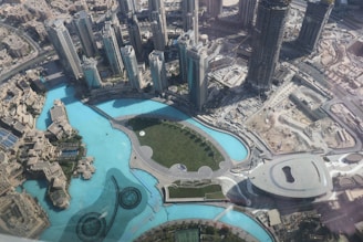 An aerial view of a modern cityscape featuring tall skyscrapers surrounding a large man-made lake with a curved promenade. The architecture is futuristic with symmetrical patterns and landscaping, and the scene also includes open spaces, roads, and construction areas.