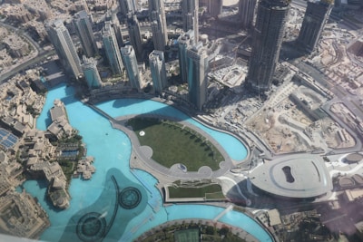 An aerial view of a modern cityscape featuring tall skyscrapers surrounding a large man-made lake with a curved promenade. The architecture is futuristic with symmetrical patterns and landscaping, and the scene also includes open spaces, roads, and construction areas.