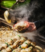 A steaming bowl of Korean street food, likely odeng, is being served. The scene includes a variety of fish cakes in a steaming tray. A person is holding a ladle and pouring broth into a red bowl filled with different shapes of fish cakes.