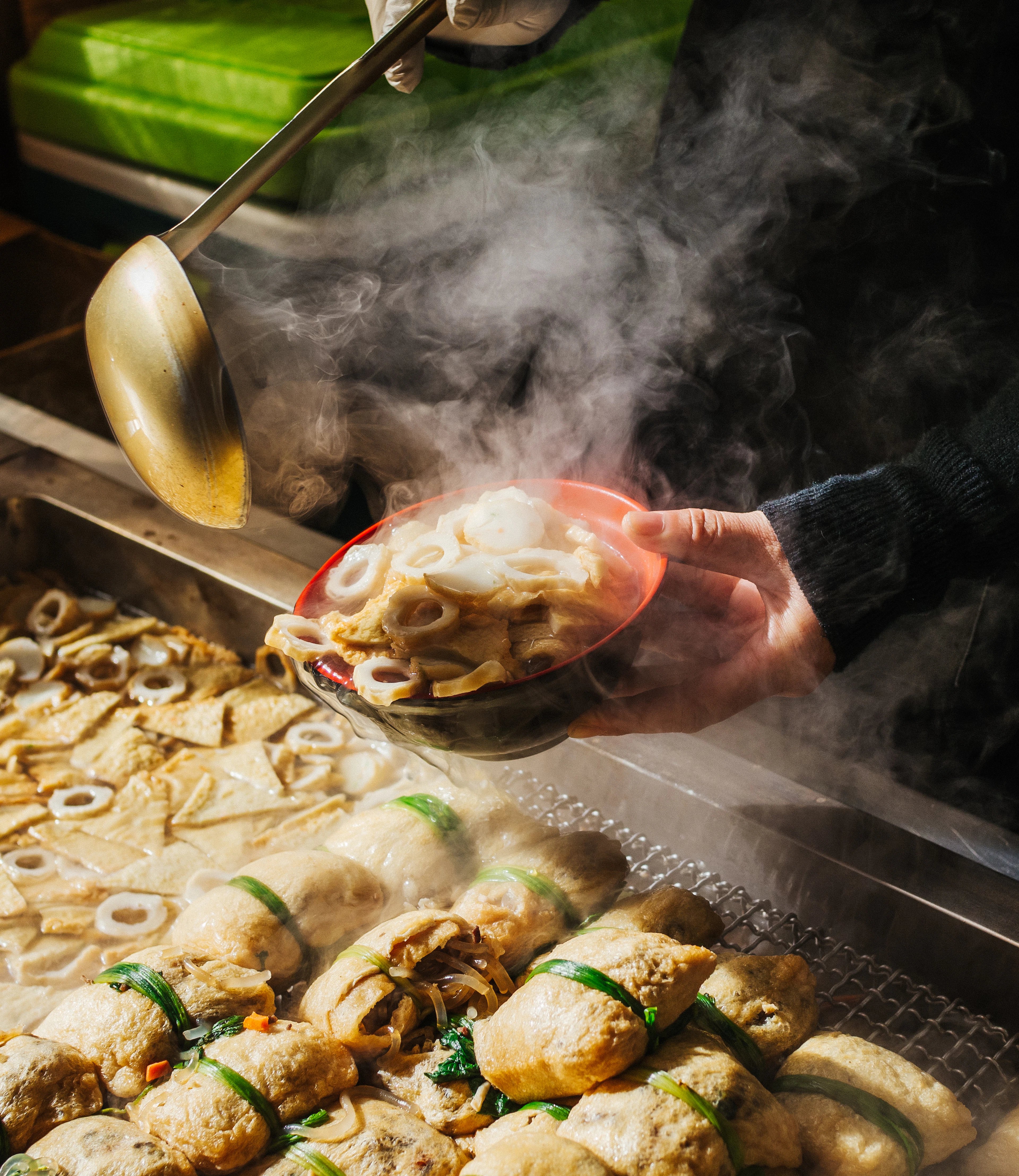 A steaming bowl of eomuk fish cake being passed across a Korean street stall, skewers of fish cake simmering in broth on the griddle below, Busan's signature snack