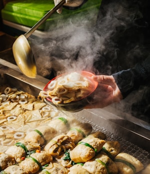 A steaming bowl of Korean street food, likely odeng, is being served. The scene includes a variety of fish cakes in a steaming tray. A person is holding a ladle and pouring broth into a red bowl filled with different shapes of fish cakes.