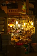 A small, bustling market stall is packed with various goods packaged in plastic bags and containers. The warm glow of multiple hanging lightbulbs illuminates the assortment of items, creating a cozy ambiance. Egg cartons and cardboard boxes are stacked in front of the stall, while dried goods hang above, catching the light.