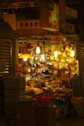 A small, bustling market stall is packed with various goods packaged in plastic bags and containers. The warm glow of multiple hanging lightbulbs illuminates the assortment of items, creating a cozy ambiance. Egg cartons and cardboard boxes are stacked in front of the stall, while dried goods hang above, catching the light.