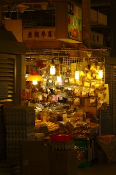 A small, bustling market stall is packed with various goods packaged in plastic bags and containers. The warm glow of multiple hanging lightbulbs illuminates the assortment of items, creating a cozy ambiance. Egg cartons and cardboard boxes are stacked in front of the stall, while dried goods hang above, catching the light.