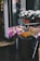 A small flower shop display featuring various bouquets and potted plants. Pink and white flowers are wrapped in vibrant pink paper, while pots of white flowers are arranged on a metal stand. Orange flowers are displayed in a dark container with a price tag. The scene is set on a rainy day with wet pavement.