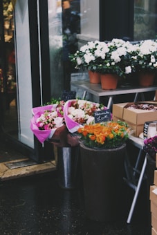 A small flower shop display featuring various bouquets and potted plants. Pink and white flowers are wrapped in vibrant pink paper, while pots of white flowers are arranged on a metal stand. Orange flowers are displayed in a dark container with a price tag. The scene is set on a rainy day with wet pavement.