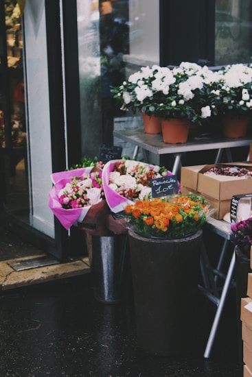 A small flower shop display featuring various bouquets and potted plants. Pink and white flowers are wrapped in vibrant pink paper, while pots of white flowers are arranged on a metal stand. Orange flowers are displayed in a dark container with a price tag. The scene is set on a rainy day with wet pavement.