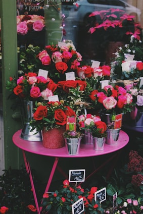 A vibrant display of colorful flowers arranged on a pink table outside a flower shop. The arrangement includes red and pink roses, placed in various pots and metal containers with price tags. The shop window reflects a car and more plant arrangements in the background.