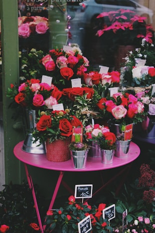 A vibrant display of colorful flowers arranged on a pink table outside a flower shop. The arrangement includes red and pink roses, placed in various pots and metal containers with price tags. The shop window reflects a car and more plant arrangements in the background.