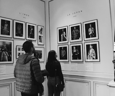 Two people stand in an art gallery observing a collection of framed photographs on the wall. The wall is divided into sections labeled 'L'INDE' for India and 'LE JAPON' for Japan, with black and white portraits showcasing traditional attire from each country. The setting features ornate wall paneling and an elegant, minimalistic design.