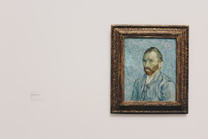 A framed painting of a man with a reddish-brown beard, wearing a blue coat and a white shirt, is displayed on a plain wall. The background of the painting features swirling blue patterns.