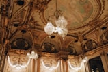 An opulent interior with intricate architectural detailing adorned with gold elements. Crystal chandeliers hang from the ceiling, casting a warm glow. The ceiling features elaborate frescoes and decorative moldings. The room exudes luxury and grandeur.