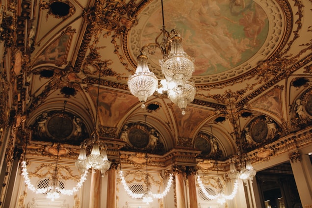 An opulent interior with intricate architectural detailing adorned with gold elements. Crystal chandeliers hang from the ceiling, casting a warm glow. The ceiling features elaborate frescoes and decorative moldings. The room exudes luxury and grandeur.