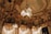 An opulent interior with intricate architectural detailing adorned with gold elements. Crystal chandeliers hang from the ceiling, casting a warm glow. The ceiling features elaborate frescoes and decorative moldings. The room exudes luxury and grandeur.