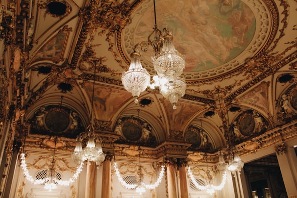 An opulent interior with intricate architectural detailing adorned with gold elements. Crystal chandeliers hang from the ceiling, casting a warm glow. The ceiling features elaborate frescoes and decorative moldings. The room exudes luxury and grandeur.