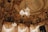 An opulent interior with intricate architectural detailing adorned with gold elements. Crystal chandeliers hang from the ceiling, casting a warm glow. The ceiling features elaborate frescoes and decorative moldings. The room exudes luxury and grandeur.