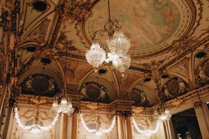 An opulent interior with intricate architectural detailing adorned with gold elements. Crystal chandeliers hang from the ceiling, casting a warm glow. The ceiling features elaborate frescoes and decorative moldings. The room exudes luxury and grandeur.