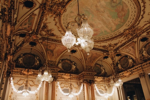 An opulent interior with intricate architectural detailing adorned with gold elements. Crystal chandeliers hang from the ceiling, casting a warm glow. The ceiling features elaborate frescoes and decorative moldings. The room exudes luxury and grandeur.