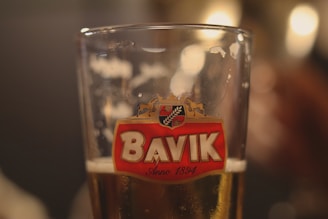 A close-up of a glass of beer with a Bavik logo prominently displayed, featuring a red emblem with a horse and hops. The glass is partially filled with a light amber beer, and the background is softly blurred, creating a warm, inviting ambiance.
