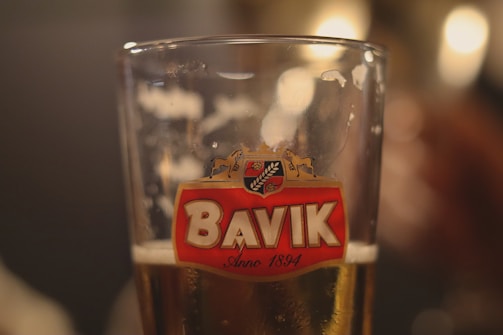 A close-up of a glass of beer with a Bavik logo prominently displayed, featuring a red emblem with a horse and hops. The glass is partially filled with a light amber beer, and the background is softly blurred, creating a warm, inviting ambiance.