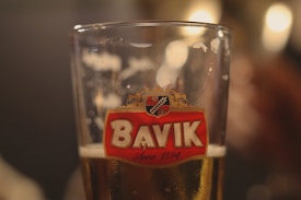 A close-up of a glass of beer with a Bavik logo prominently displayed, featuring a red emblem with a horse and hops. The glass is partially filled with a light amber beer, and the background is softly blurred, creating a warm, inviting ambiance.