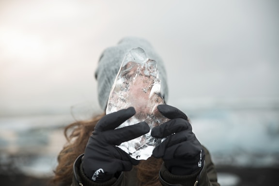 A person is holding a large, clear piece of ice up to their face with both hands. The person is wearing gloves and a beanie, and the background is a blurred wintry landscape with an overcast sky.