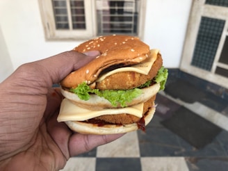 A hand holding a large sandwich with multiple layers, including sesame seed buns, cheese slices, lettuce, and breaded patties. The setting appears to be indoors with white walls and a tiled floor visible in the background.