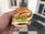 A hand holding a large sandwich with multiple layers, including sesame seed buns, cheese slices, lettuce, and breaded patties. The setting appears to be indoors with white walls and a tiled floor visible in the background.