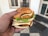 A hand holding a large sandwich with multiple layers, including sesame seed buns, cheese slices, lettuce, and breaded patties. The setting appears to be indoors with white walls and a tiled floor visible in the background.
