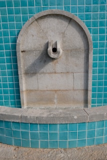 A stone fountain is set against a wall covered with teal ceramic tiles. The fountain has an arched top and a protruding spout in the center. The stone appears weathered, suggesting age and exposure to the elements.