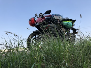 A motorcycle packed with gear stands amidst tall green grass, positioned against a clear blue sky. The red helmet attached to the motorcycle adds a pop of color, and the vehicle appears ready for a journey or adventure.