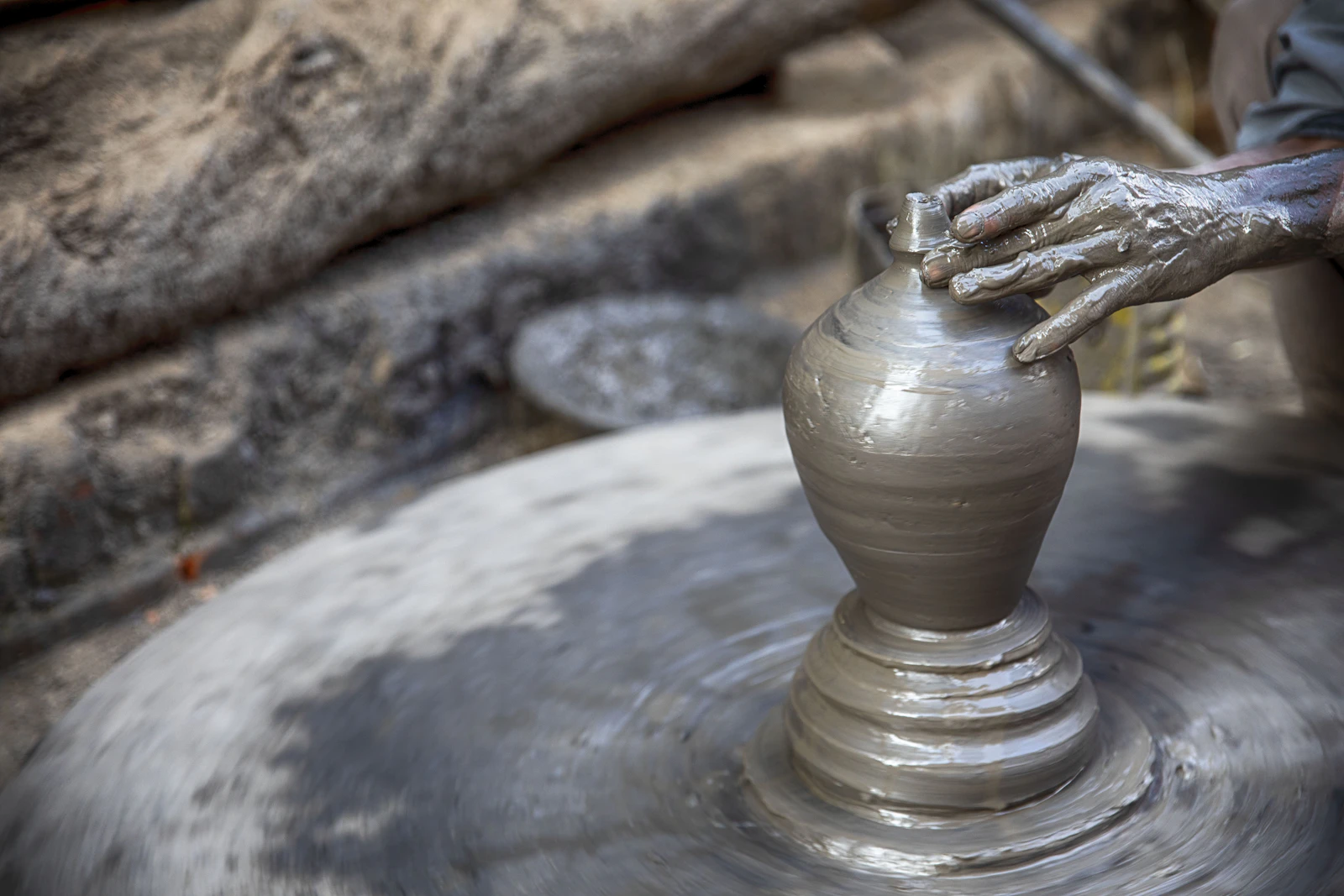 A potter's clay-covered hands shaping a grey pot on a spinning wheel in Pottery Square, Bhaktapur