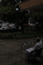 An elderly person in white clothing is seated on a chair outdoors, near a walking frame. The surrounding area includes a concrete patio, potted plants, and trees. In the background, there is a street with vehicles and buildings, all partially obscured by foliage.