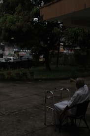 An elderly person in white clothing is seated on a chair outdoors, near a walking frame. The surrounding area includes a concrete patio, potted plants, and trees. In the background, there is a street with vehicles and buildings, all partially obscured by foliage.