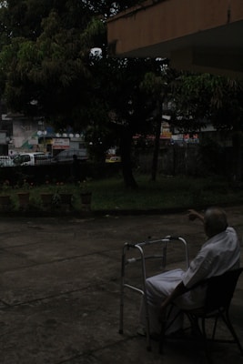 An elderly person in white clothing is seated on a chair outdoors, near a walking frame. The surrounding area includes a concrete patio, potted plants, and trees. In the background, there is a street with vehicles and buildings, all partially obscured by foliage.