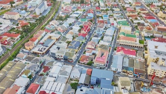 Aerial view of a densely packed urban neighborhood with a mix of low to mid-rise buildings and colorful rooftops. The streets are arranged in a grid pattern, and there is a visible train track running parallel to one side. The buildings vary in architectural style, with several modern structures mixed with older ones, and many rooftops are painted in shades of red, blue, and green.