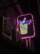 A vibrant neon sign depicting a relaxed character leaning back in a chair, holding a drink. The sign features bright colors with a pink outline, set against a nighttime urban backdrop. Adjacent air conditioning units and wires are visible, suggesting a city environment.