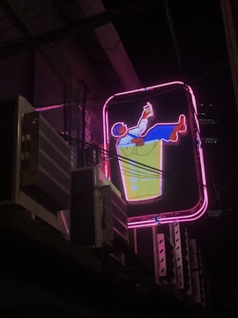 A vibrant neon sign depicting a relaxed character leaning back in a chair, holding a drink. The sign features bright colors with a pink outline, set against a nighttime urban backdrop. Adjacent air conditioning units and wires are visible, suggesting a city environment.
