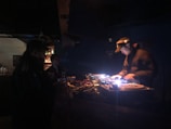 A dimly lit nightclub scene featuring a DJ operating a mixing console with colorful lights. There are visible turntables and a crowd in the background. The ambiance is dark with scattered vibrant lights reflecting off the equipment.
