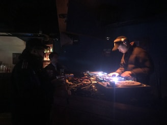 A dimly lit nightclub scene featuring a DJ operating a mixing console with colorful lights. There are visible turntables and a crowd in the background. The ambiance is dark with scattered vibrant lights reflecting off the equipment.