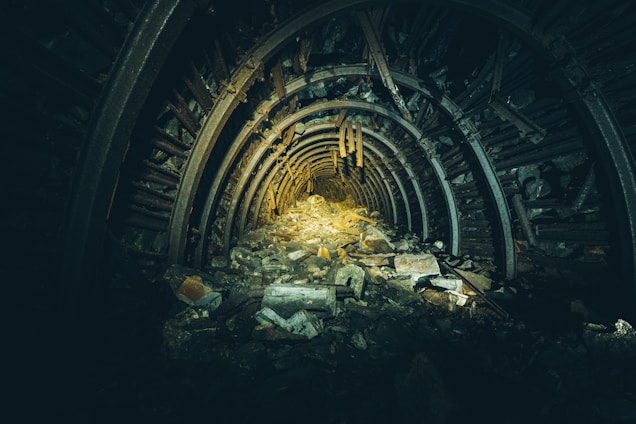 An abandoned underground tunnel featuring a rugged, debris-strewn path. The tunnel is framed by concentric metal supports, with the far end dimly lit, emphasizing the desolation and disarray. The lighting creates contrasting shadows and highlights the tunnel's worn, rusty texture.